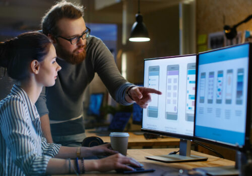 Female UX Architect Has Discussion with Male Design Engineer, They Work on Mobile Application Late at Night. In the Background Wall with Project Sticky Notes and Other Studio Employees.